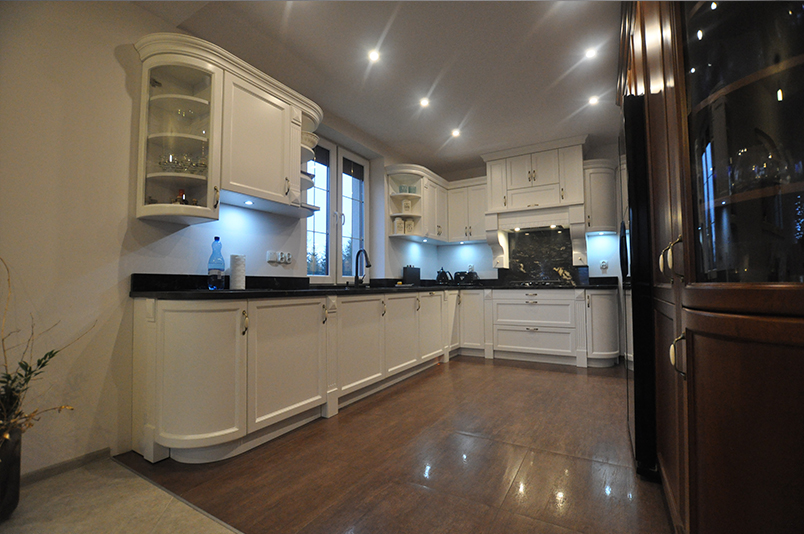 Klasyczna biała kuchnia z giętymi szafkami, czarnym granitowym blatem, witryną szklaną i oświetleniem LED.

