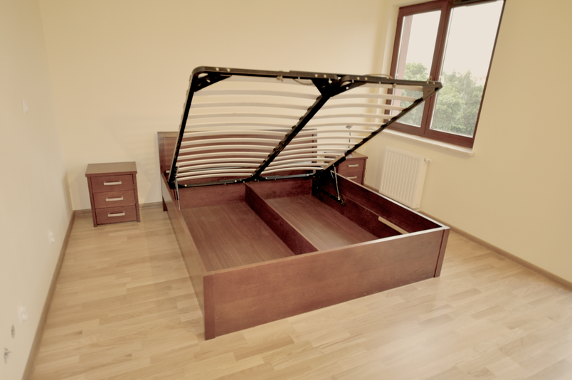 Drewniane łóżko mahoniowe z podnoszonym stelażem i pojemnikiem na pościel, otwierane do góry w jasnej sypialni z szafką nocną.